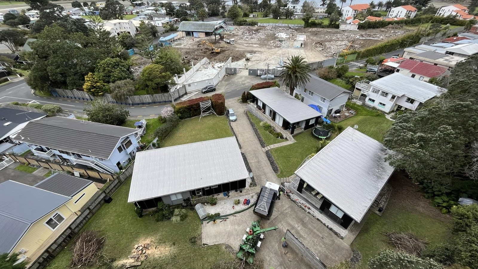 Aerial view over a seaside street of houses towards the beach and an offshore island, a green chipper and cut timber in a back yard in the foreground
