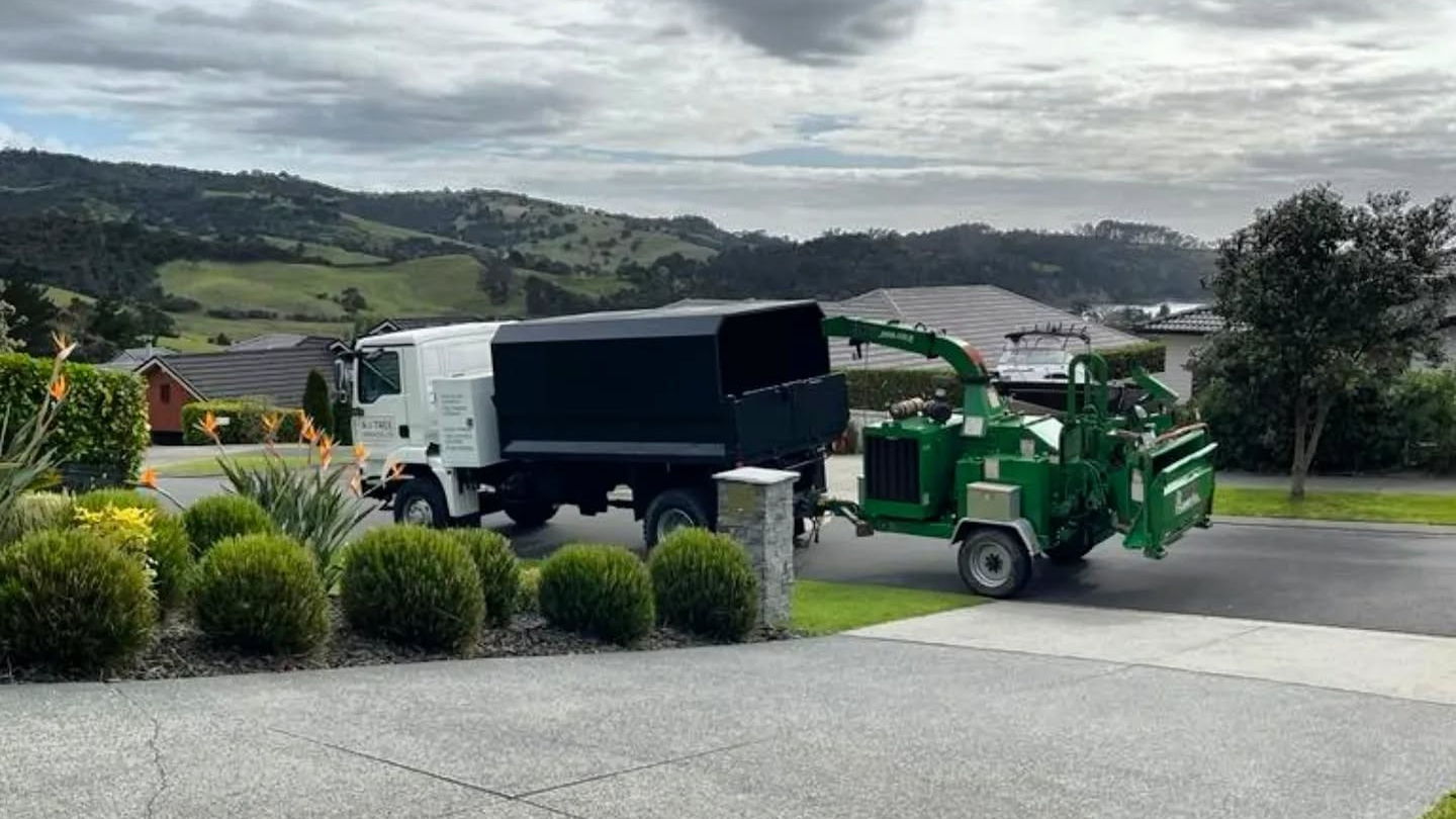 An A To Tree tipper truck and green wood chipper parked on a concrete driveway, with green hills behind