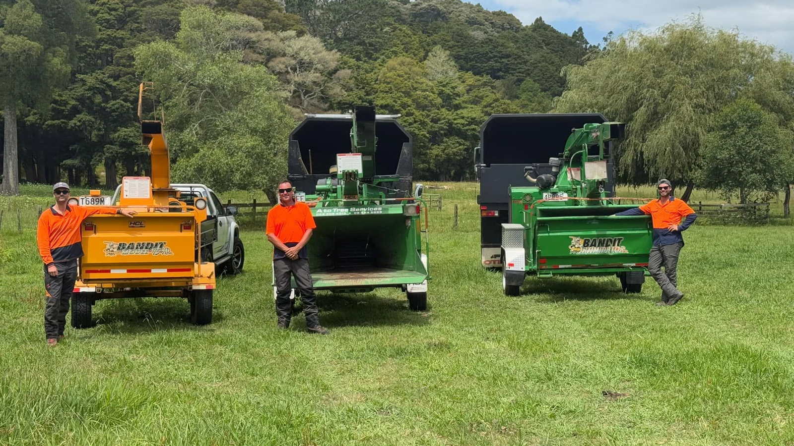 Three A To Tree crew members in orange hi-vis standing with their three Bandit wood chippers, one orange and two green, in a grass paddock with bush-covered hills behind