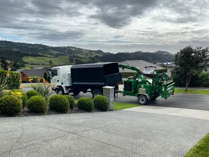 An A To Tree tipper truck and green wood chipper parked on a concrete driveway, with green hills behind