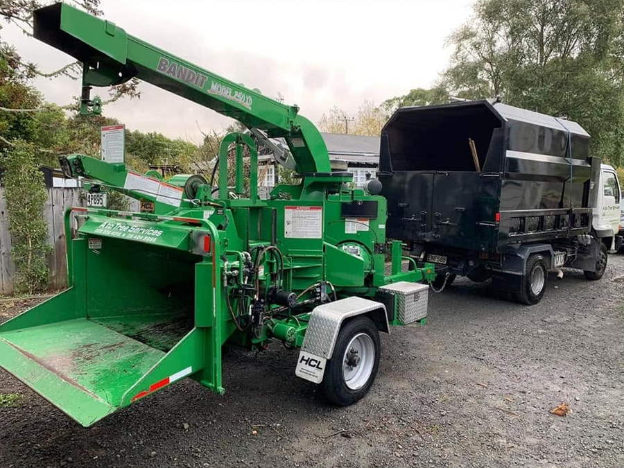A green Bandit wood chipper hitched to a black-bodied tipper truck on a gravel drive