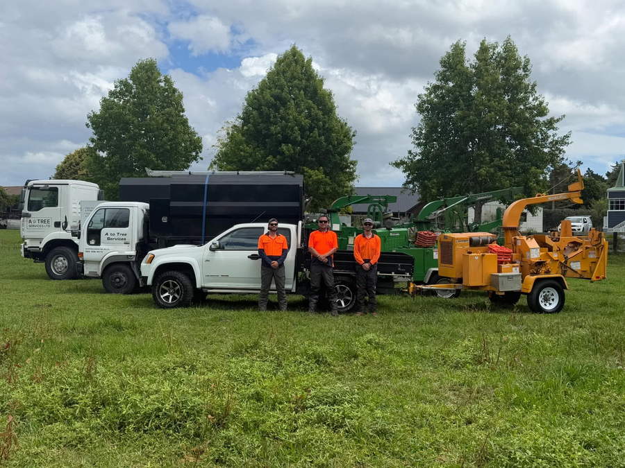 The A To Tree crew in orange hi-vis in front of their two tipper trucks, a white ute and two wood chippers, parked on grass under tall trees