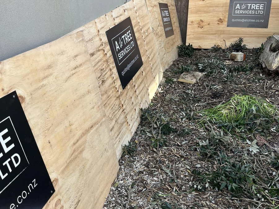Plywood protection boards carrying A To Tree Services signs along a wall, with a bed of fresh wood chip mulch in front