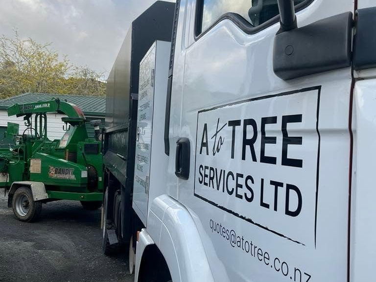 The door of an A To Tree Services Ltd truck, signwritten with quotes@atotree.co.nz, a green chipper parked behind