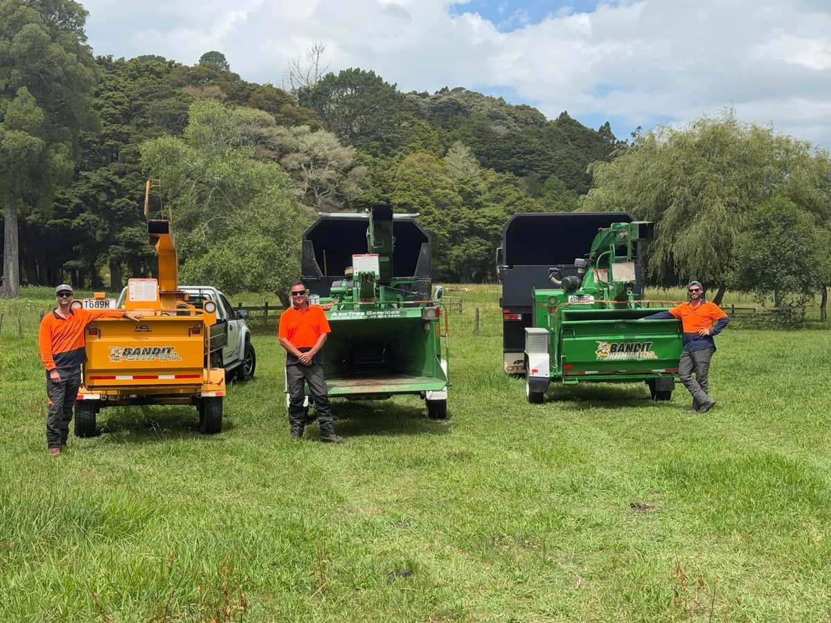 Three A To Tree crew members in orange hi-vis standing with their three Bandit wood chippers, one orange and two green, in a grass paddock with bush-covered hills behind
